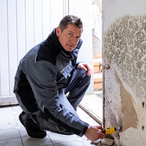 Mitarbeiter in Arbeitsjacke arbeitet in der Hocke an einer stark verschimmelten Wand neben der Tür